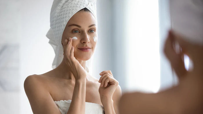Woman applying cream to her face with a towel on her head, looking at herself in the mirror.