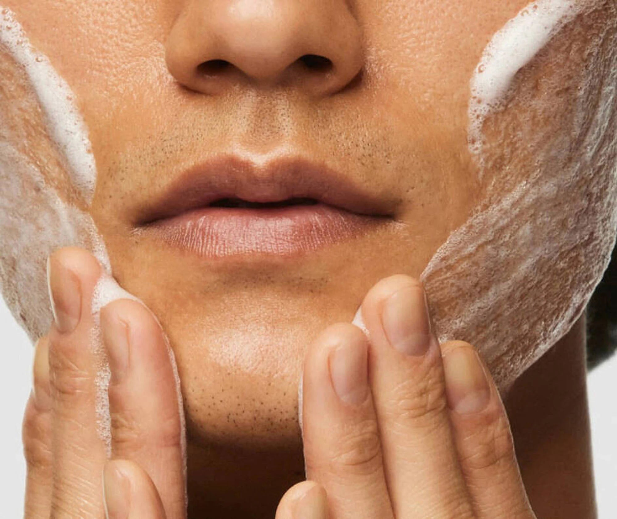 Close-up of a person applying cleanser to their face with a neutral background