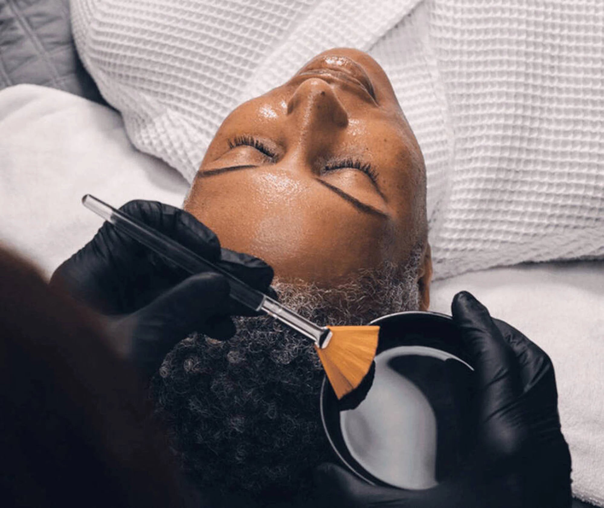 Person receiving a facial treatment with a brush and mask in a spa setting.