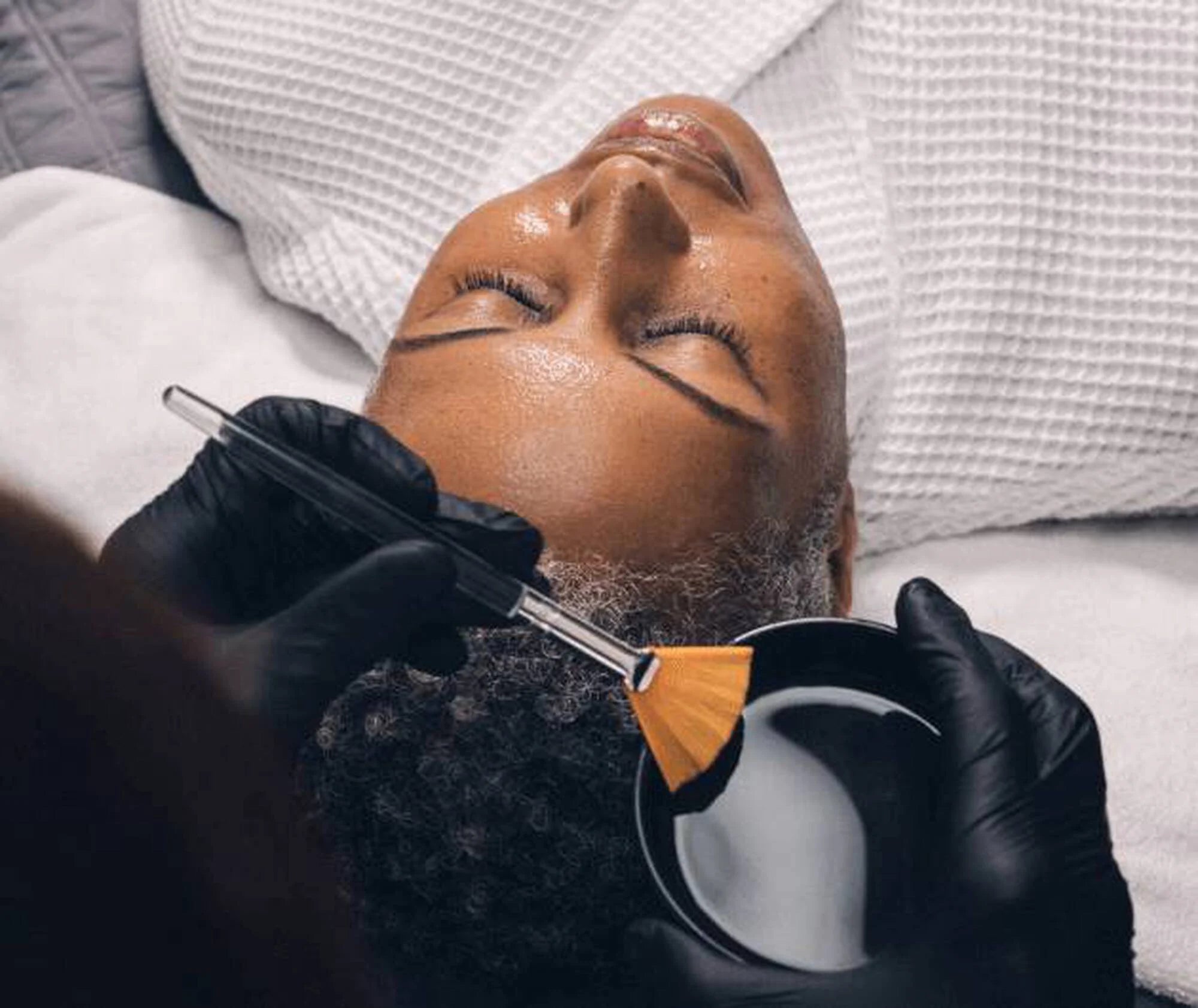 Person receiving a facial treatment with a brush and container of product.