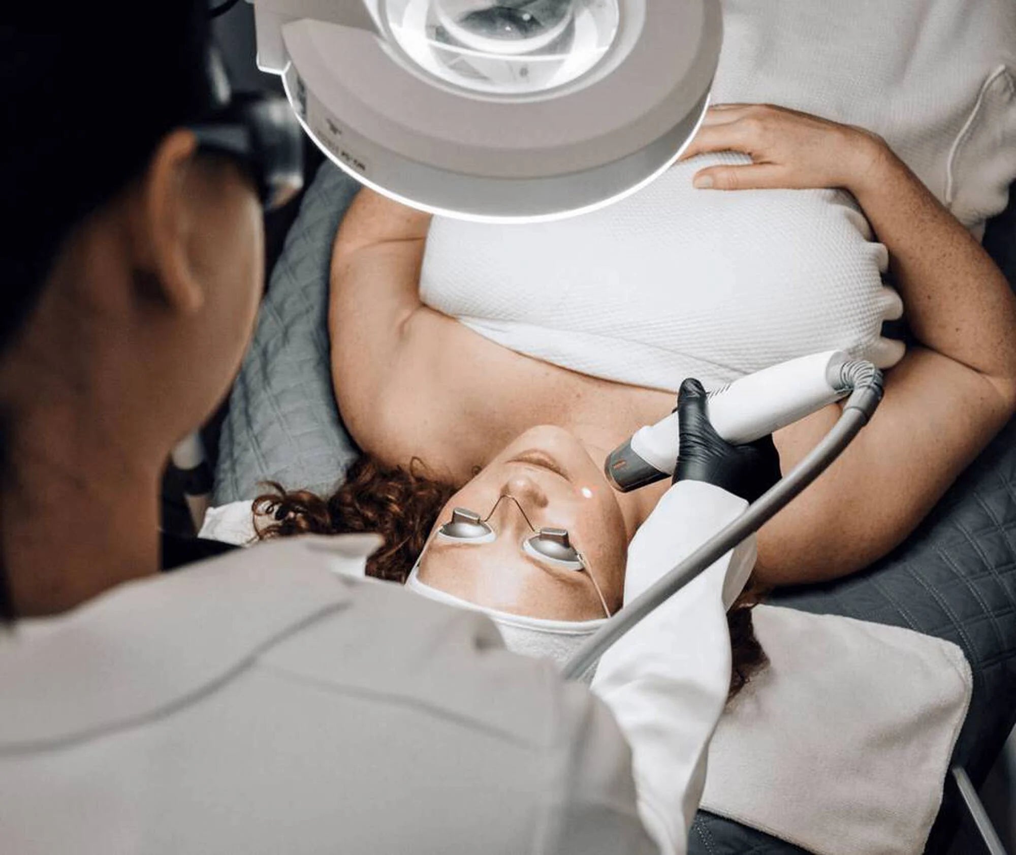 Person receiving a cosmetic procedure under a lamp with medical equipment.
