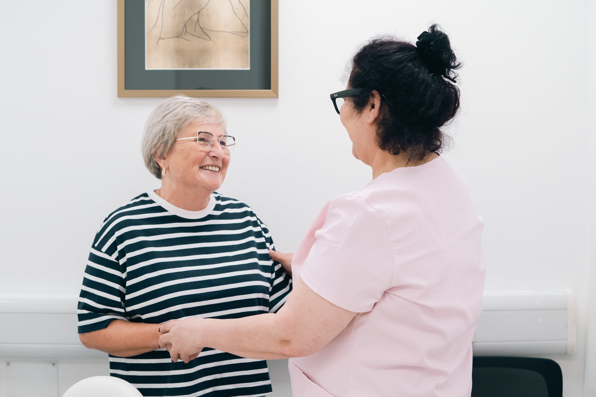 Two women shaking hands in a room with a framed picture on the wall.