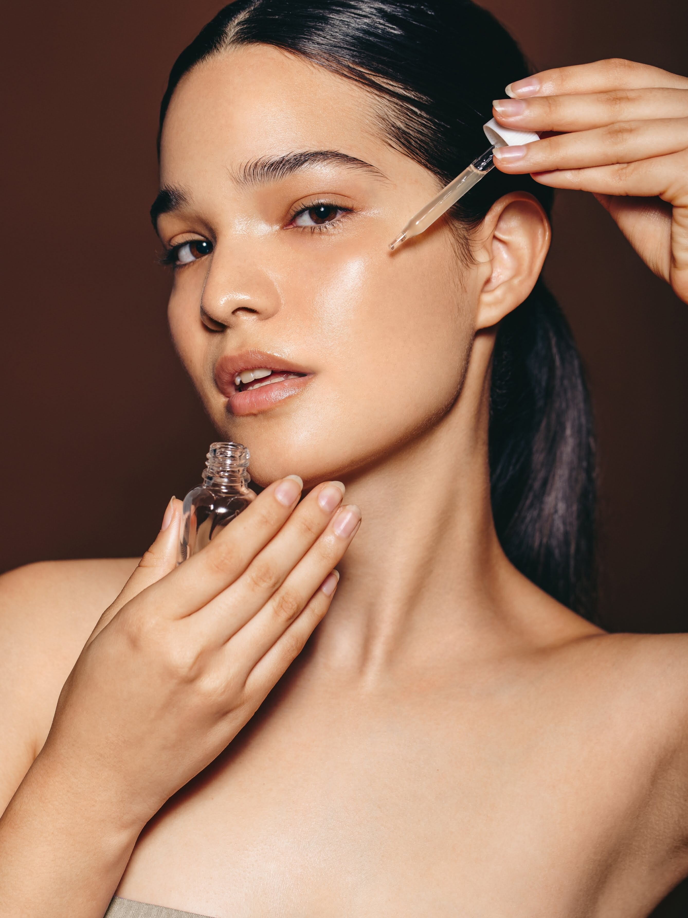 Woman applying skincare product with dropper to face against a brown background